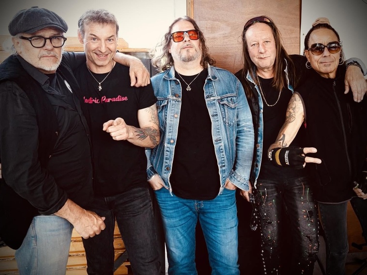 Five men in rock band attire pose together, some with denim jackets, sunglasses, and tattoos, smiling at the camera (group portrait).