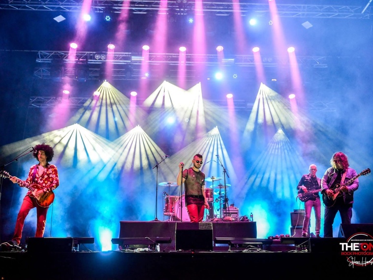 Live rock band on stage performing under colorful purple and blue lights with a singer center stage and guitarists on sides.