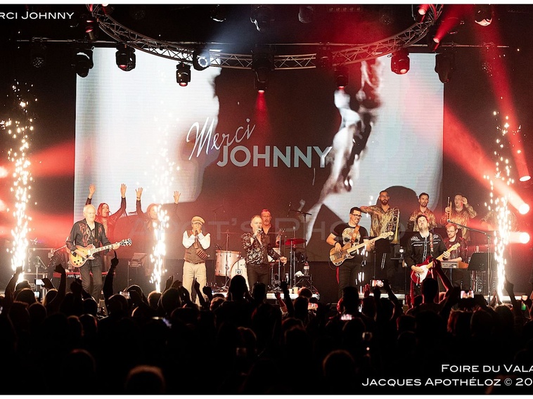 Live concert on a large stage with a multi-member band performing, sparks fly from the sides as a cheering crowd watches, a screen reads 'Merci Johnny'.