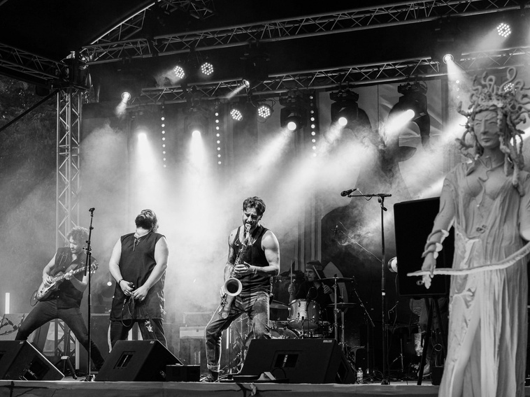Black-and-white photo of a four-piece rock band performing on an outdoor stage with dramatic lights and a statue on the right.