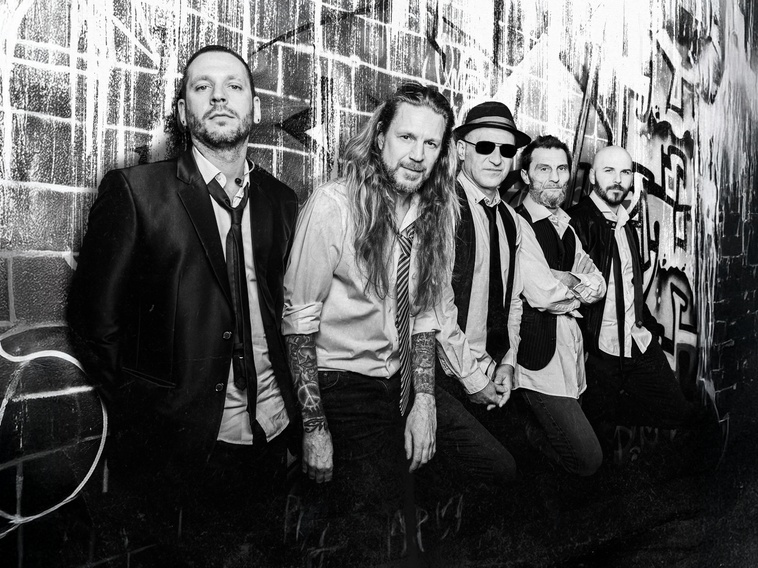 Six male musicians pose against a graffiti-covered wall, wearing shirts, ties, and vests; visible tattoos on arms.
